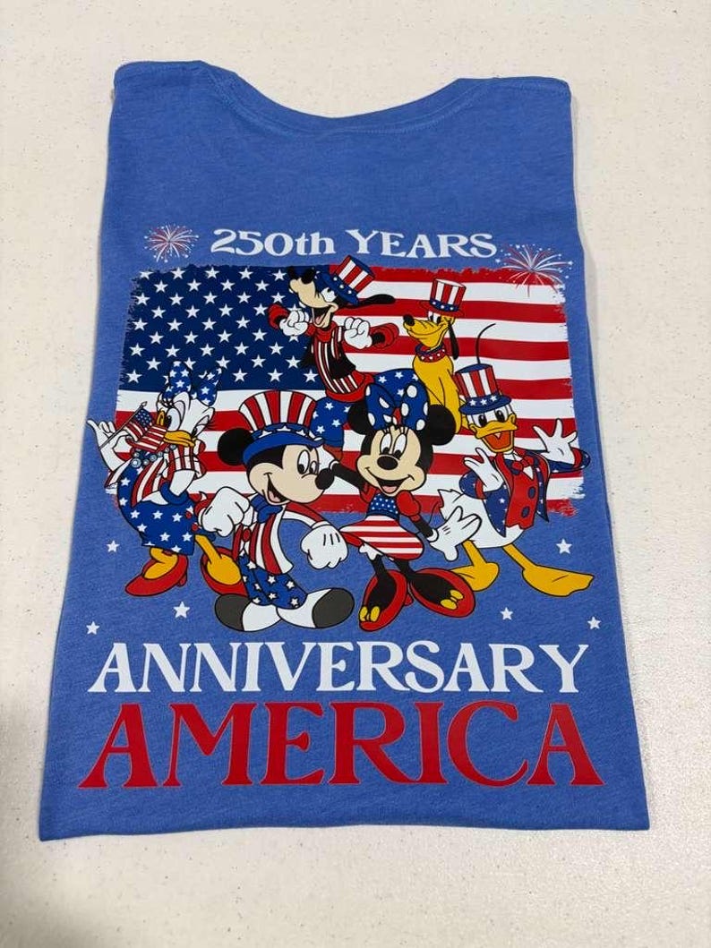 May include: A blue t-shirt featuring a graphic of Disney characters wearing patriotic red, white, and blue outfits in front of an American flag. The text reads 250th YEARS ANNIVERSARY AMERICA, accompanied by firework illustrations at the top corners.