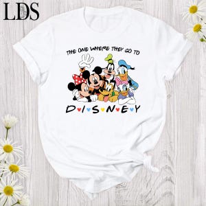 Puede incluir: Camiseta blanca con un dise&ntilde;o de Disney que presenta a Mickey y Minnie Mouse, Goofy, Donald y Daisy Duck. El texto dice "THE ONE WHERE THEY GO TO DISNEY" y "DISNEY" con corazones. La camiseta tiene mangas enrolladas.