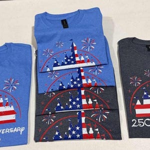 May include: Light blue and dark gray t-shirts featuring a graphic of a castle silhouette filled with a red, white, and blue American flag pattern, surrounded by fireworks. The text reads 250th Anniversary 2026 with a Mickey Mouse head icon.