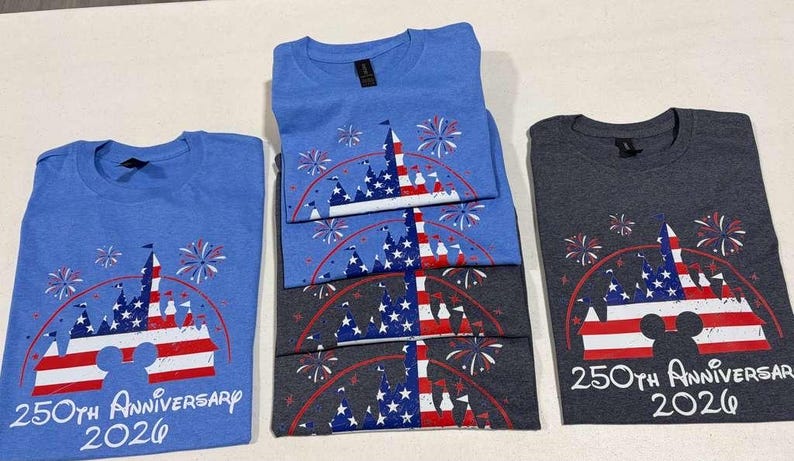 May include: Light blue and dark gray t-shirts featuring a graphic of a castle silhouette filled with a red, white, and blue American flag pattern, surrounded by fireworks. The text reads 250th Anniversary 2026 with a Mickey Mouse head icon.