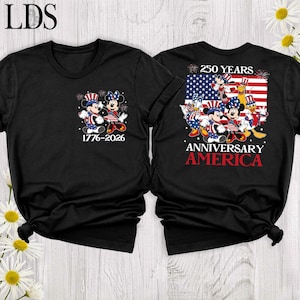 May include: A black t-shirt featuring a front graphic of Mickey and Minnie Mouse in patriotic attire with the text 1776-2026. The back displays a large American flag with Disney characters, the text 250 YEARS ANNIVERSARY AMERICA, and fireworks.