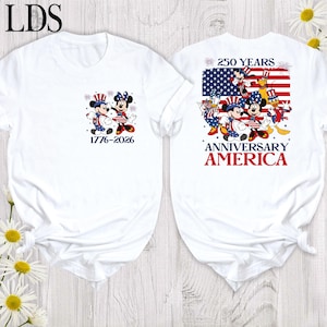 May include: A white t-shirt featuring a small graphic on the front with Mickey and Minnie Mouse and the text 1776-2026. The back displays a large American flag graphic with Disney characters, the text 250 YEARS ANNIVERSARY AMERICA, and fireworks.