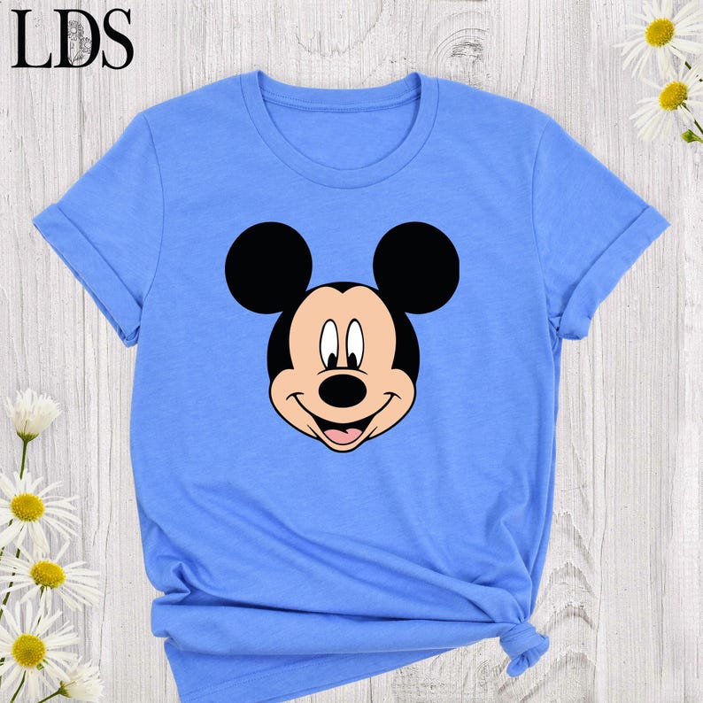 May include: Light blue t-shirt featuring a large graphic of Mickey Mouse's face. The cartoon character's face is in black, white, and tan. The shirt has short sleeves and a crew neck. The letters "LDS" are in the upper left corner.