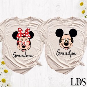Puede incluir: Dos camisetas beige con gr&aacute;ficos de personajes de dibujos animados. Una presenta a Minnie Mouse con un lazo rojo y gafas, la palabra "Grandma" debajo. La otra tiene a Mickey Mouse con gafas y la palabra "Grandpa".