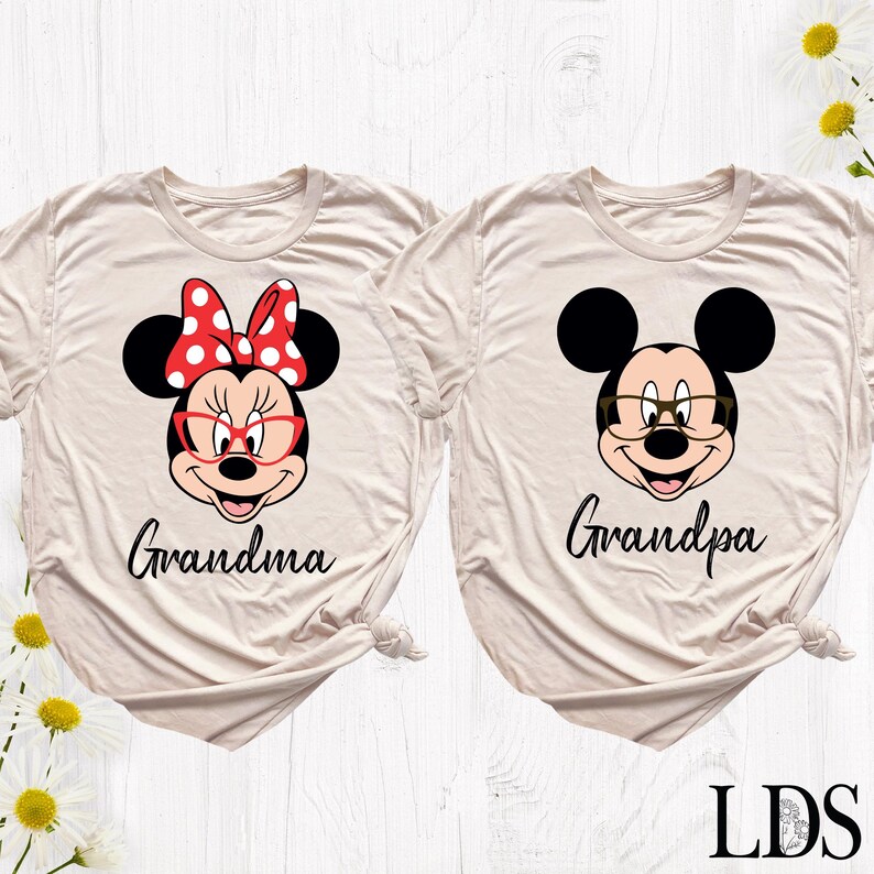 Puede incluir: Dos camisetas beige con gr&aacute;ficos de personajes de dibujos animados. Una presenta a Minnie Mouse con un lazo rojo y gafas, la palabra "Grandma" debajo. La otra tiene a Mickey Mouse con gafas y la palabra "Grandpa".