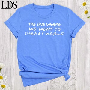 May include: A light blue short-sleeved t-shirt featuring the white text "THE ONE WHERE WE WENT TO DISNEY WORLD" printed on the front. The letters in "DISNEY" are separated by small, colorful dots in red, blue, yellow, and green hues.