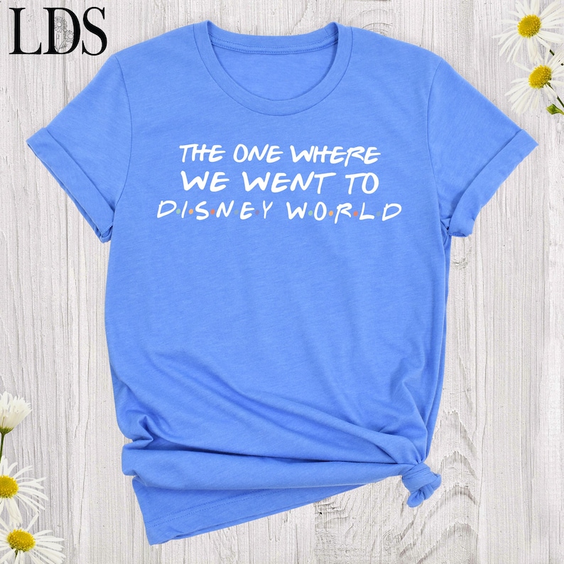 May include: A light blue short-sleeved t-shirt featuring the white text "THE ONE WHERE WE WENT TO DISNEY WORLD" printed on the front. The letters in "DISNEY" are separated by small, colorful dots in red, blue, yellow, and green hues.