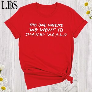 May include: A bright red short-sleeved t-shirt featuring white text that reads THE ONE WHERE WE WENT TO DISNEY WORLD, with small colored dots separating the letters in the word DISNEY and the word WORLD, and a crew neckline.