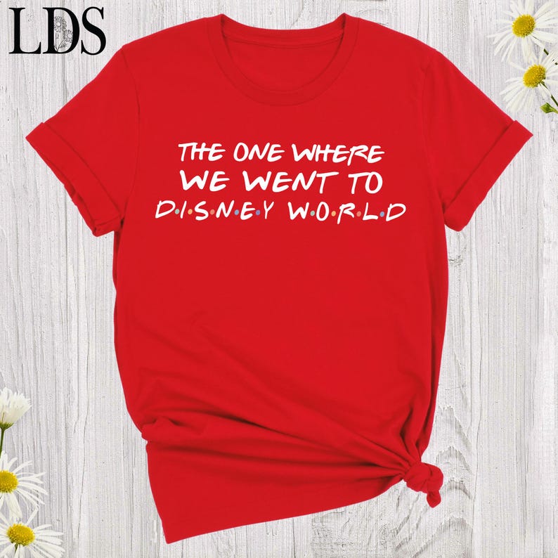 May include: A bright red short-sleeved t-shirt featuring white text that reads THE ONE WHERE WE WENT TO DISNEY WORLD, with small colored dots separating the letters in the word DISNEY and the word WORLD, and a crew neckline.
