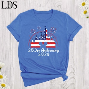 May include: A blue short-sleeve t-shirt featuring a graphic of a castle silhouette filled with a red, white, and blue American flag pattern. Above the castle are fireworks, and below is the text 250th Anniversary 2026 in a white script font.