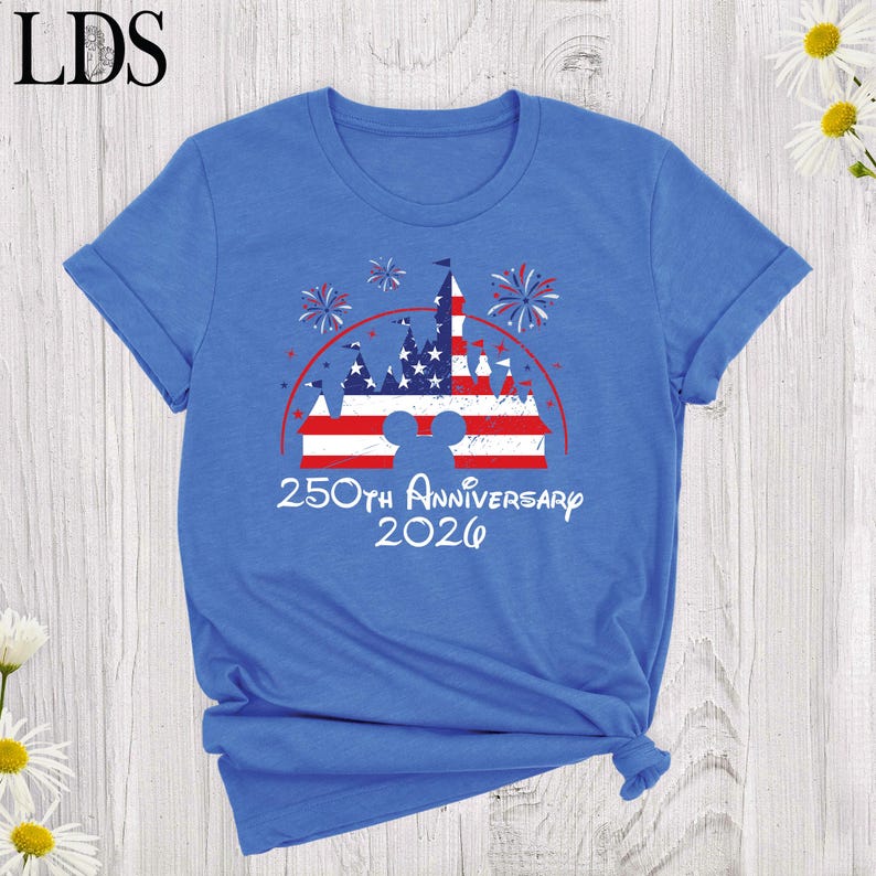 May include: A blue short-sleeve t-shirt featuring a graphic of a castle silhouette filled with a red, white, and blue American flag pattern. Above the castle are fireworks, and below is the text 250th Anniversary 2026 in a white script font.
