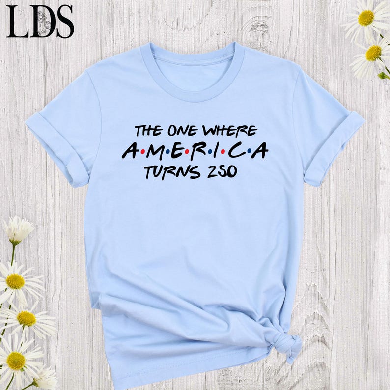 May include: A light blue t-shirt with rolled-up sleeves. The shirt features the text "THE ONE WHERE AMERICA TURNS 250" in black and red lettering, reminiscent of a popular sitcom's logo. The shirt is made of a soft material.