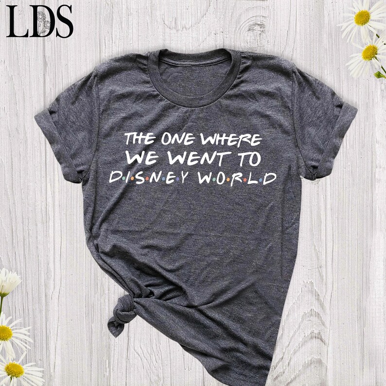 May include: A dark heather gray short-sleeve t-shirt with a crew neckline. The front features white text that reads THE ONE WHERE WE WENT TO DISNEY WORLD, with small colored dots separating the letters in the word DISNEY.