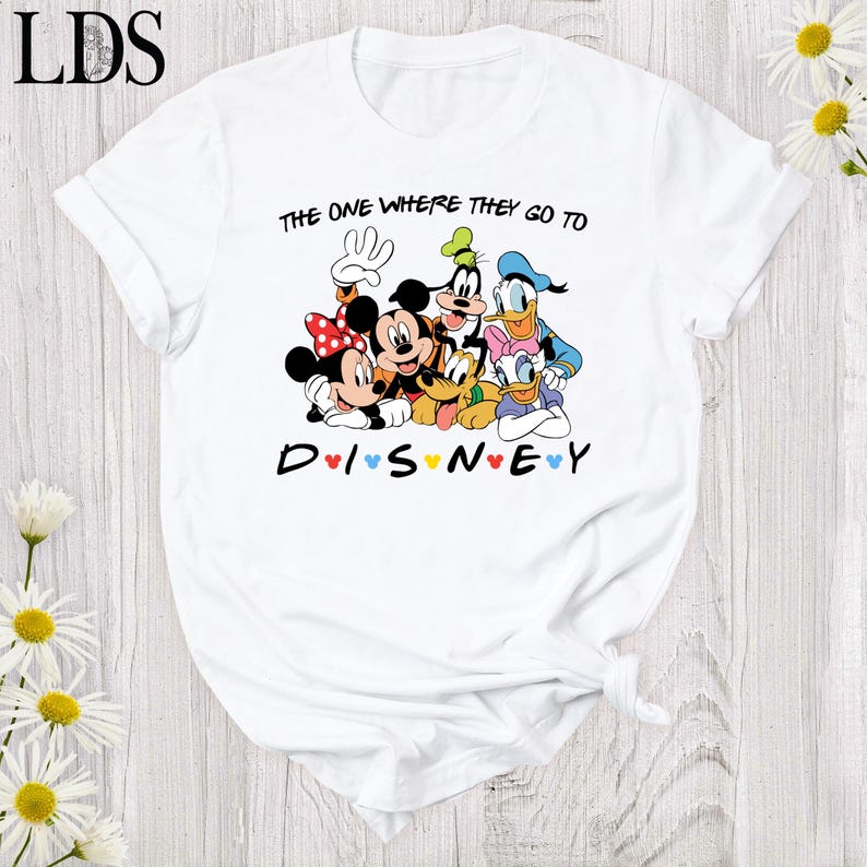 Puede incluir: Camiseta blanca con un dise&ntilde;o de Disney que presenta a Mickey y Minnie Mouse, Goofy, Donald y Daisy Duck. El texto dice "THE ONE WHERE THEY GO TO DISNEY" y "DISNEY" con corazones. La camiseta tiene mangas enrolladas.