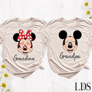 Puede incluir: Dos camisetas beige con gr&aacute;ficos de personajes de dibujos animados. Una presenta a Minnie Mouse con un lazo rojo y gafas, la palabra "Grandma" debajo. La otra tiene a Mickey Mouse con gafas y la palabra "Grandpa".