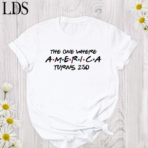 May include: White t-shirt with the text "THE ONE WHERE AMERICA TURNS 250" in black and red lettering. The design is reminiscent of the Friends TV show logo. The shirt is made of a soft material and has short sleeves.