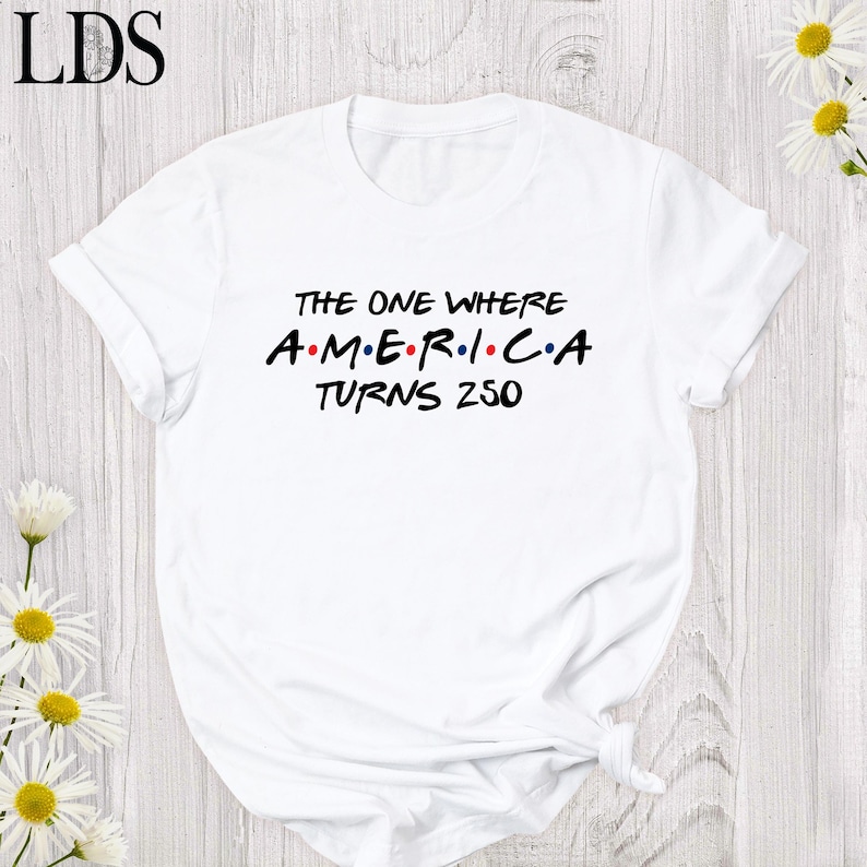 May include: White t-shirt with the text "THE ONE WHERE AMERICA TURNS 250" in black and red lettering. The design is reminiscent of the Friends TV show logo. The shirt is made of a soft material and has short sleeves.