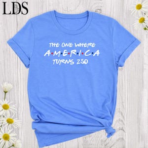 May include: A light blue t-shirt with rolled-up sleeves. The shirt features the text "THE ONE WHERE A.M.E.R.I.C.A TURNS 250" in white and red letters. The shirt is tied at the bottom.