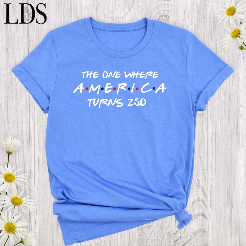 May include: A light blue t-shirt with rolled-up sleeves. The shirt features the text "THE ONE WHERE A.M.E.R.I.C.A TURNS 250" in white and red letters. The shirt is tied at the bottom.