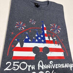 May include: A charcoal gray t-shirt featuring a graphic of a castle silhouette filled with a red, white, and blue American flag pattern. Above the castle are red and blue fireworks, and below is the white text 250TH ANNIVERSARY 2026.