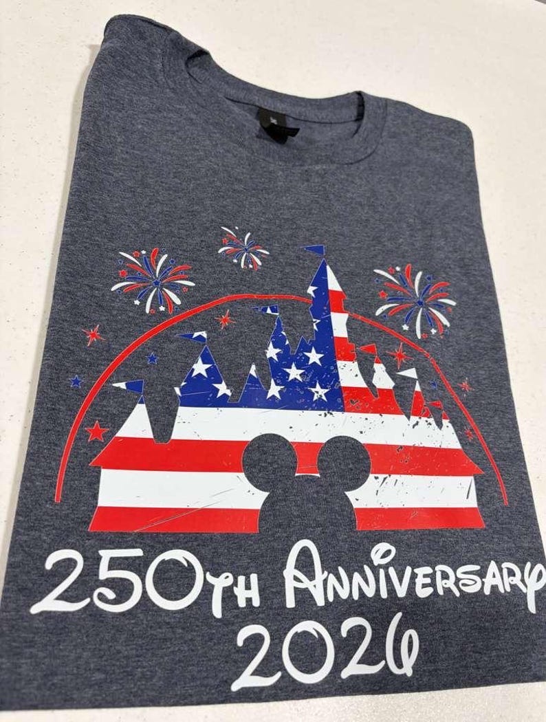 May include: A charcoal gray t-shirt featuring a graphic of a castle silhouette filled with a red, white, and blue American flag pattern. Above the castle are red and blue fireworks, and below is the white text 250TH ANNIVERSARY 2026.