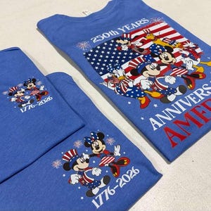 May include: Two blue t-shirts featuring Disney characters in patriotic outfits. The larger shirt displays a flag graphic with the text 250th YEARS ANNIVERSARY AMERICA. The smaller shirts feature Mickey and Minnie Mouse with the text 1776-2026.
