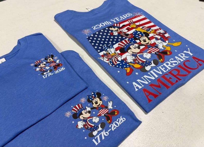 May include: Two blue t-shirts featuring Disney characters in patriotic outfits. The larger shirt displays a flag graphic with the text 250th YEARS ANNIVERSARY AMERICA. The smaller shirts feature Mickey and Minnie Mouse with the text 1776-2026.
