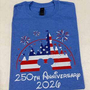 May include: A blue short-sleeve t-shirt featuring a graphic of a castle silhouette filled with a red, white, and blue American flag pattern, surrounded by fireworks, with the text 250th Anniversary 2026 printed in white below the castle.