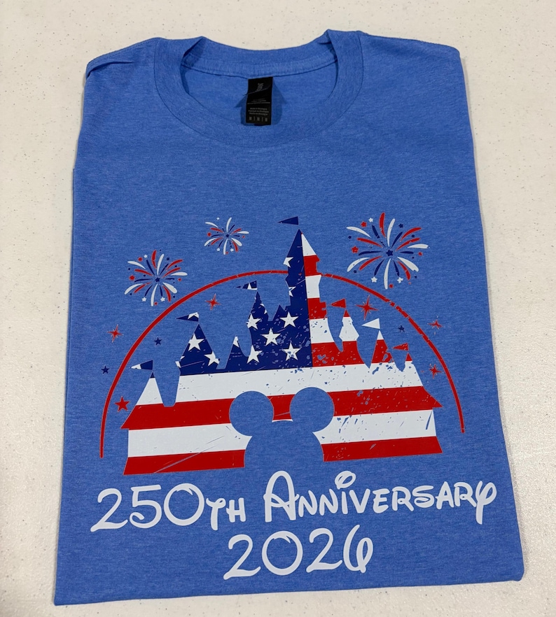 May include: A blue short-sleeve t-shirt featuring a graphic of a castle silhouette filled with a red, white, and blue American flag pattern, surrounded by fireworks, with the text 250th Anniversary 2026 printed in white below the castle.