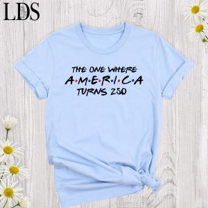 May include: A light blue t-shirt with rolled-up sleeves. The shirt features the text "THE ONE WHERE AMERICA TURNS 250" in black and red lettering, reminiscent of a popular sitcom's logo. The shirt is made of a soft material.