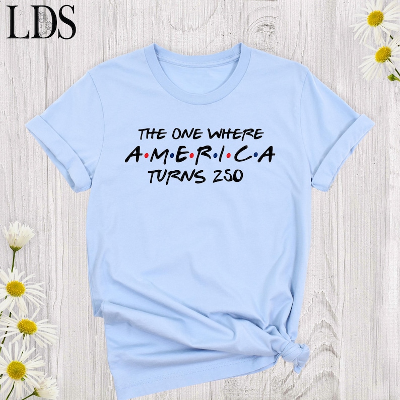 May include: A light blue t-shirt with rolled-up sleeves. The shirt features the text "THE ONE WHERE AMERICA TURNS 250" in black and red lettering, reminiscent of a popular sitcom's logo. The shirt is made of a soft material.