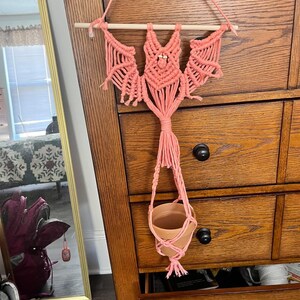 Pode incluir: Suporte de planta em macramé feito à mão na cor rosa coral. O design tem formato de morcego e é pendurado em uma vareta de madeira. A cesta tecida suporta um pequeno vaso de planta marrom claro.