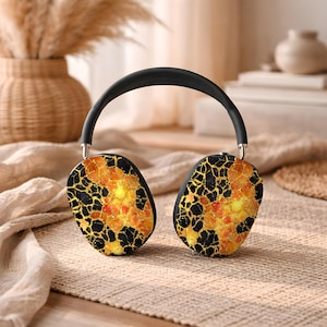 May include: Black and gold wireless headphones with a unique mosaic design. The earcups feature a pattern of black lines and gold and orange accents. The headband is black, and the headphones are resting on a beige surface.