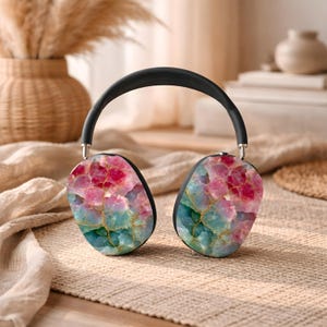 May include: Black over-ear headphones with earcups featuring a colorful, abstract design in shades of pink, red, teal, and gold. The headband is black, and the earcups have a smooth, rounded shape. The headphones are resting on a neutral-toned surface.