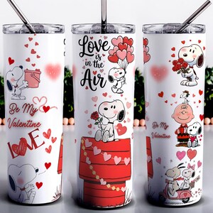 May include: White stainless steel tumbler featuring Snoopy and Woodstock Valentine's Day designs. The tumbler has a silver lid and straw. Designs include Snoopy with hearts, the text "Love is in the Air", and "Be My Valentine".