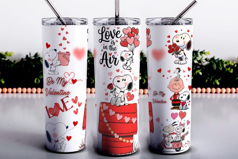 May include: White stainless steel tumbler featuring Snoopy and Woodstock Valentine's Day designs. The tumbler has a silver lid and straw. Designs include Snoopy with hearts, the text "Love is in the Air", and "Be My Valentine".