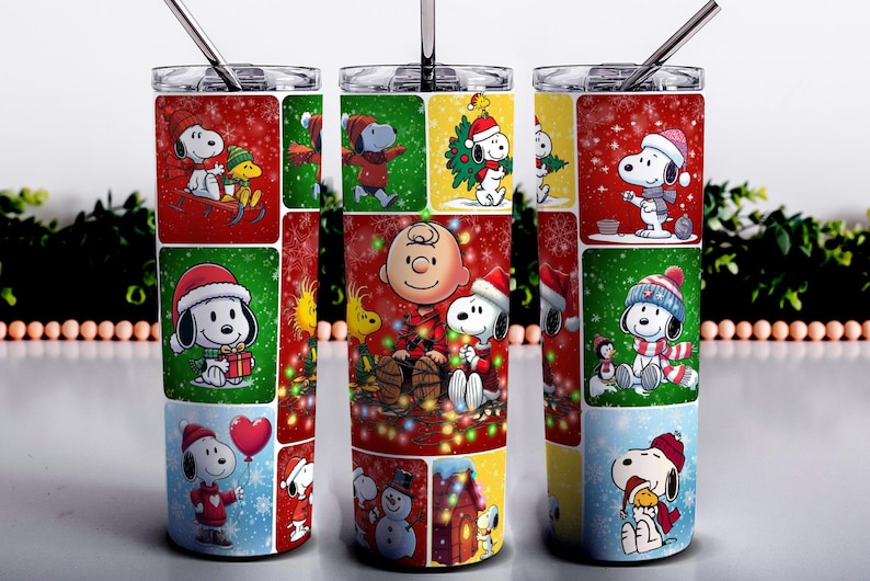 May include: A stainless steel tumbler featuring a festive design with Snoopy and Charlie Brown characters in a Christmas theme. The tumbler has a straw and a lid, and the design includes red, green, and blue colors.