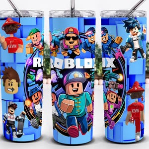 May include: A blue tumbler with a Roblox theme, showcasing various character illustrations. The design incorporates the Roblox logo and several game characters in action poses. The tumbler has a silver straw and a clear lid.