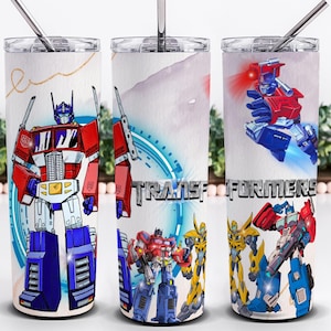 May include: Three white stainless steel tumblers with illustrations of Transformers characters. The tumblers feature Optimus Prime in red, white, and blue, and the word "TRANSFORMERS" in silver. Each tumbler has a silver straw and a white lid.