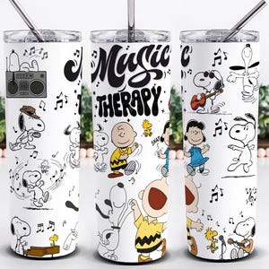 以下が含まれることがあります： スヌーピーと仲間たちが踊ったり音楽を演奏したりする、銀色のストローが付いた白いタンブラー。「Music Therapy」の文字が黒の様式化されたフォントで表示されています。音符とブームボックスもデザインに含まれています。
