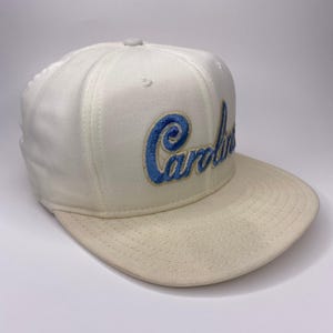 K&ouml;nnte beinhalten: Cremefarbene Baseballkappe mit gebogenem Schirm. Das Wort "Carolina" ist in blauer Schrift auf die Vorderseite gestickt. Die Kappe hat einen Knopf oben und eine strukturierte Krone.