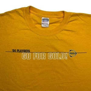 May include: Mustard yellow t-shirt with the text "'04 PLAYOFFS GO FOR GOLD!" in white and black. The shirt has a crew neck and a small tag on the upper center. A logo is on the right side of the text.