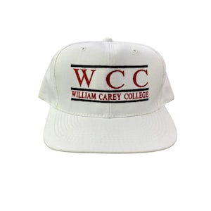May include: White baseball cap with a flat brim. The front of the cap features the letters "WCC" in red, with "WILLIAM CAREY COLLEGE" embroidered below in red. The text is framed by two black horizontal lines.