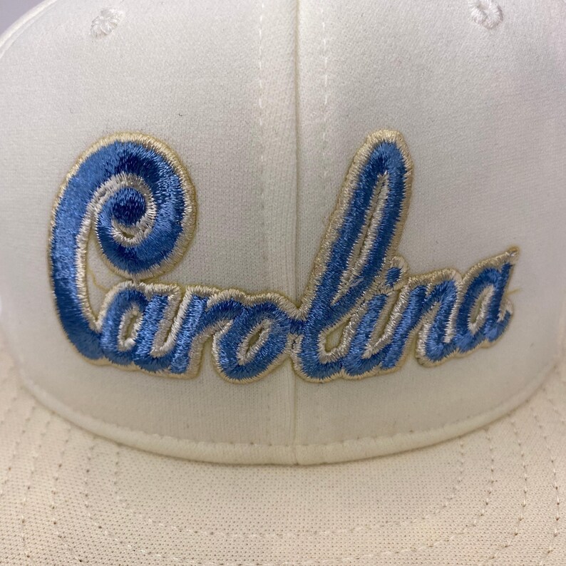 K&ouml;nnte beinhalten: Cremefarbene Baseballkappe mit dem Wort "Carolina" in hellblauer Schrift mit goldener Umrandung gestickt. Die Kappe hat eine strukturierte Krone und einen gebogenen Schirm, geeignet f&uuml;r Sonnenschutz.