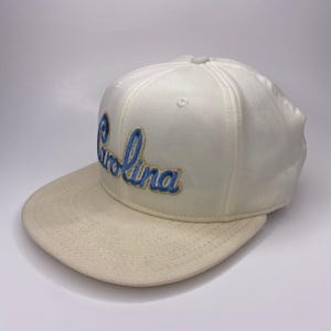 K&ouml;nnte beinhalten: Cremefarbene Baseballkappe mit flachem Schirm. Das Wort "Carolina" ist in blauer Schrift auf der Vorderseite gestickt. Die Kappe hat eine strukturierte Krone und einen Knopf oben.