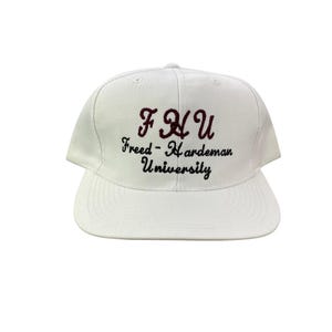 Puede incluir: Gorra de béisbol blanca con visera plana. La gorra presenta el texto bordado "FHU" en granate, sobre las palabras "Freed-Hardeman University" en escritura negra. Una gorra de estilo clásico.
