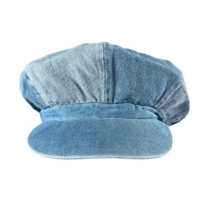 May include: A newsboy cap crafted from light blue denim fabric. The hat features a structured, rounded crown with visible vertical seams and a stiff, curved brim. The denim material displays a washed-out texture with variations in blue tones.