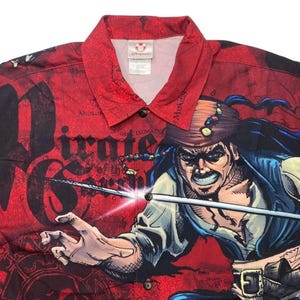 May include: Red button-up shirt featuring a graphic of Captain Jack Sparrow from Pirates of the Caribbean. The shirt has a repeating pattern of text and imagery, with a Disney tag at the collar.