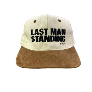 May include: A vintage baseball cap with a tan crown and a brown suede brim. The text "LAST MAN STANDING" is printed in bold black letters across the front. The cap has a classic six-panel design.