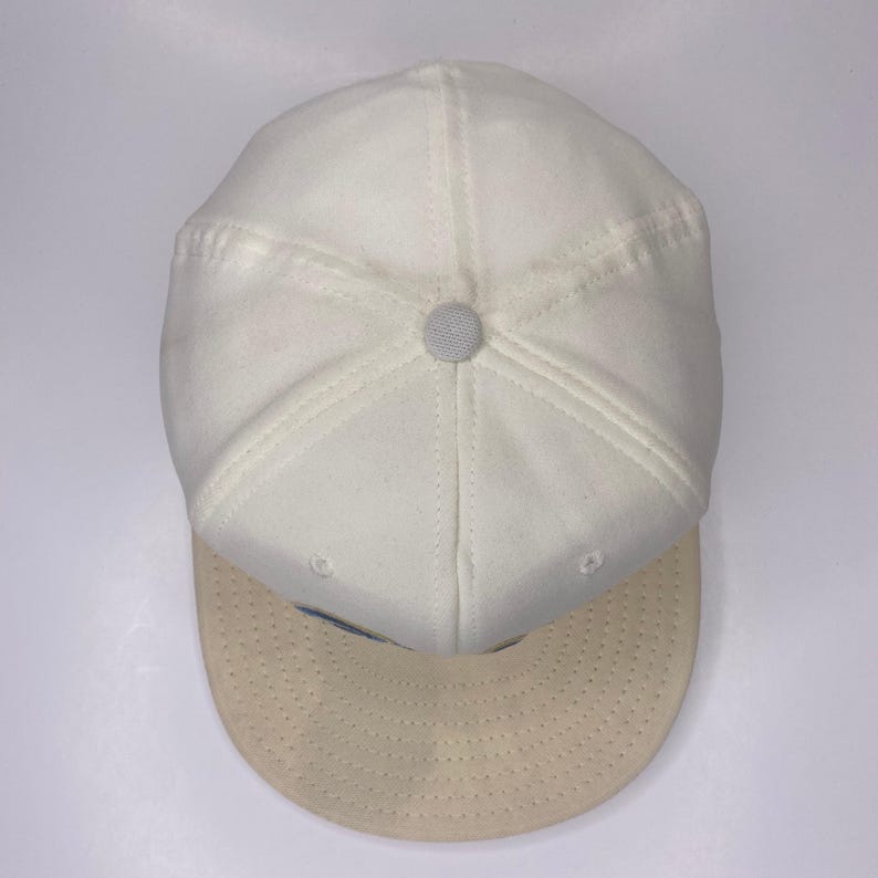 K&ouml;nnte beinhalten: Eine wei&szlig;e Baseballkappe mit hellbeigem Schirm. Die Kappe hat einen Knopf oben und Nahtdetails. Der Schirm ist ein hellerer Beigeton. Die Kappe wird von oben betrachtet.